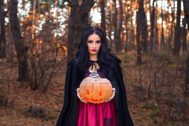 Zarif siyah elbiseli gizemli bir kadın büyülü, büyüleyici bir ormanda oyulmuş bir balkabağı tutuyor.