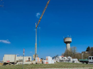 Açık mavi gökyüzüne karşı vinç, malzeme ve su kulesinin yer aldığı bir şantiye manzarası.