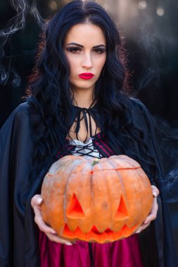 Gizemli bir ormanın derinliklerinde bir cadı oyulmuş bir balkabağını sıkıca kavrayıp ürkütücü ama büyüleyici bir atmosfer yaratıyor.