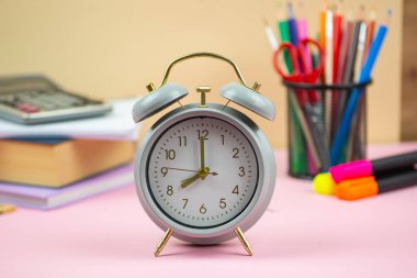 Canlı okul malzemeleri, çeşitli kitaplar ve renkli kırtasiye malzemelerinin arasına yerleştirilmiş klasik bir çalar saat.