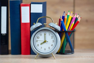 Çalar saatin, düzgünce istiflenmiş kitapların ve renkli kalemlerin sergilendiği güzel bir masa sahnesi.