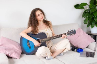 Genç bir kadın, modern estetik bir koltukta rahat rahat otururken gitar çalmaktan zevk alıyor.
