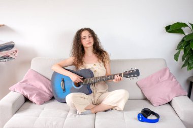 Genç bir kadın rahat bir koltukta otururken akustik gitarını çalmaktan mutlu olur.