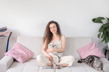 Huzurlu ve huzurlu bir an. Güzel kedilerine sevgi dolu bir şekilde sarılan bir kadının şık bir kanepede oturduğu.