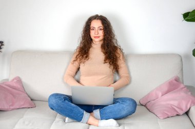 Bir kadın sıcak bir oturma odasının atmosferinde rahat bir kanepede otururken dizüstü bilgisayarı üzerinde özenle çalışıyor.