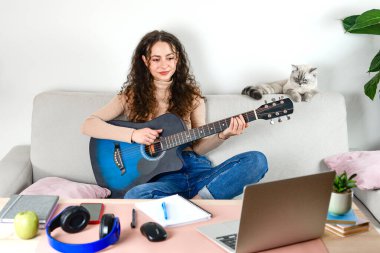 Genç bir kadın evinde gitar çalmaktan zevk alıyor, teknolojiyle dolu sıcak bir atmosferde.