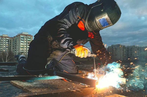 A welder in a suit and mask welds metal against the sky, welding work, a lot of sparks