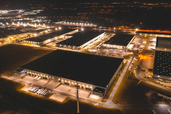 Aerial view of a warehouse of goods at night. Many warehouses and trucks with trailers are in the parking lot, waiting to be loaded for further distribution of goods around the country.