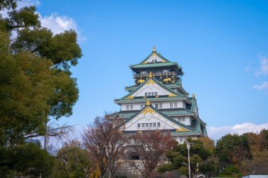 Osaka Kalesi, Japonya 'nın Osaka şehrinde açık mavi bir gökyüzünün altında görkemli bir şekilde duruyor. Kalenin geleneksel Japon mimarisi, tarihsel önemini ve güzelliğini vurgulayan çıplak ağaç dalları tarafından çerçevelenmiştir..