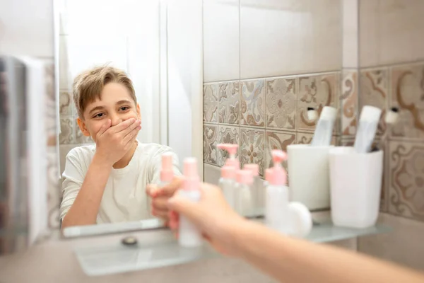 Teenager boy looking at the mirror, smiling and touching his face and holding cosmetic bottle in bathroom. Beautiful healthy child at home doing his morning skincare routine and having fun.