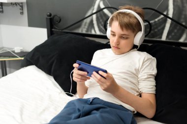Genç çocuk kulaklık takıyor, akıllı telefonla yatağa uzanıyor, oyun oynuyor ya da video seyrediyor.
