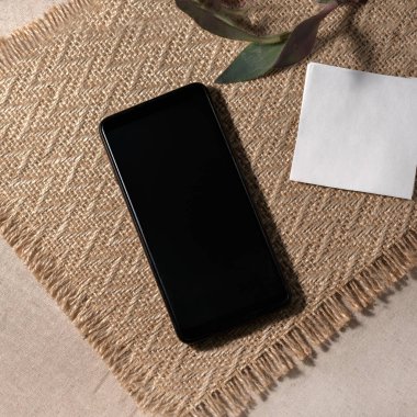 Mobile phone and blank paper sheet with mockup copy space and plant on a sand jute placemat background, minimalist aesthetic business brand template, flat lay, top view