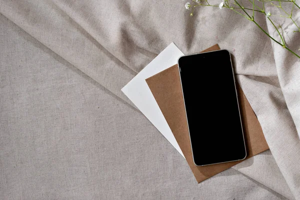 Boş siyah ekranlı cep telefonu ve nötr bej katmanlı kumaş arka plan kağıtları, fotokopi alanı ile minimalist ticari markalaşma şablonu, küçük iş yeri çalışma alanı