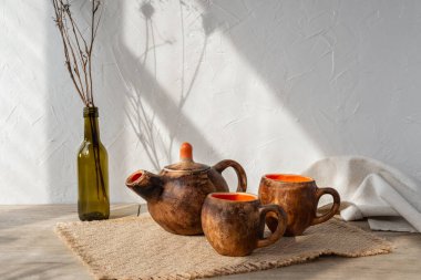 Estetik el işi çömlek çay seti, kahverengi kil çaydanlık ve kupalar ahşap bir masada, güneş ışığı gölgeleri beyaz duvarda, minimalist çay seremonisi, wabi sabi konsepti
