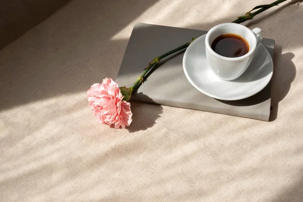 Minimalist estetik yaz durgun hayat, kahve fincanı, not defteri ve karanfil çiçeği soyut yaşam tarzı güneş ışığı gölgeleriyle bej bir masada.