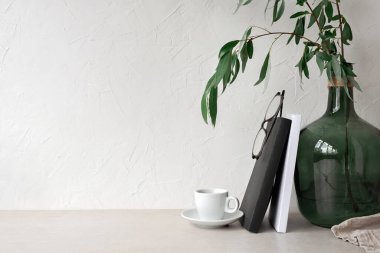 Boş nötr bej duvar fotokopi alanı arka planı. Kahve fincanı, kitaplar, bardaklar, masa üstünde bitki olan yeşil cam vazo. Minimalist modern iç mekan şablonu, ev ofis çalışma alanı, İskandinav tarzı
