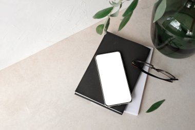 Boş cep telefonu ekran modeli, kitaplar, bardaklar, masa üstünde yeşil dalı olan vazo, boş nötr bej duvar arka planı. Minimalist ticari marka şablonu, kopyalama alanı