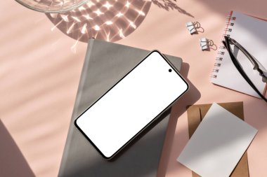 Boş akıllı telefon ekranı maketi, defter, kağıt kartlar, güneş ışığı gölgesi olan pastel pembe arka planda gözlük. Minimalist estetik kırtasiye düz döşeme, ticari marka şablonu, kopyalama alanı