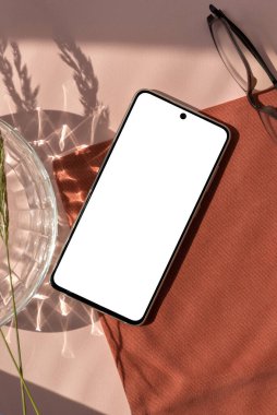 Boş cep telefonu ekranı maketi, gözlükler, estetik kristal ve çiçekli güneş ışığı gölgesi Terracotta peçetesi ve nötr şeftali bej arka plan, zarif marka şablonu
