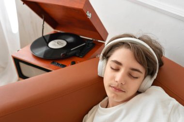 Gözleri kapalı, kulaklık takan, retro ahşap bir pikapta plaklar dinleyen, kahverengi kanepeye uzanmış, rüya gören, müzik sesinden zevk alan yakışıklı bir genç çocuk.