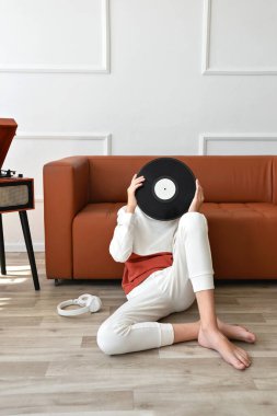 Kahverengi kanepe ve ahşap döner tablonun yanında yerde oturan genç çocuk ellerinde plak plağı tutuyordu. Yüzünü plak plağının arkasına saklıyordu. Retro vinil müzik eğilimi estetik.
