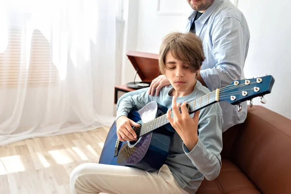 Teenager boy learning to playing an acoustic guitar with father support, sitting on sofa, at home. Fatherhood, single parent family concept. Guitar music playing classes, lifestyle.