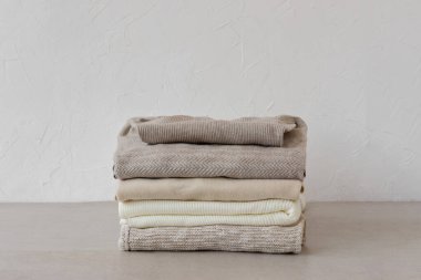 Minimalist nötr pastel bej rengi yün, kaşmir örülmüş kazaklar boş beyaz duvar arka planında bej bir masaya istiflenmiş. Sonbahar, sonbahar moda kadın kıyafetleri, örgü giysiler.