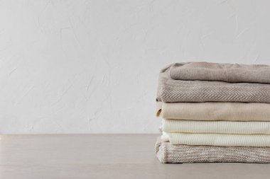 Yün örülmüş giysiler beyaz duvar arka planındaki masada nötr bej renklerde üst üste dizilir. Estetik moda minimalist kıyafet konsepti, sıcak sonbahar örgü forması