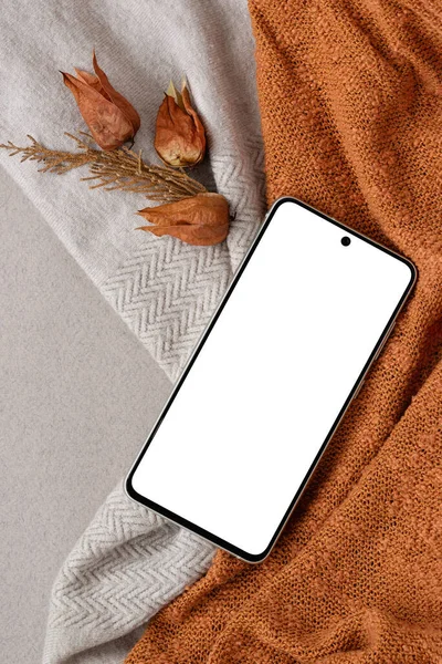 Empty mobile phone screen mock up on a neutral beige and ginger orange knitted textile background, brown dried flowers. Aesthetic business brand autumn design template, flat lay, copy space