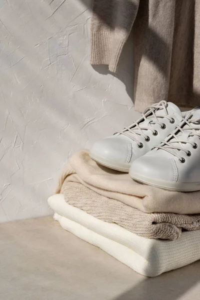 Minimalist kadın moda kıyafetleri konsepti. Tarafsız renklerde yün kazaklar, bej masadaki spor ayakkabılar ve güneş ışığı gölgeli beyaz duvar arkaplanı..