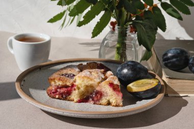 Bir parça erikli erikli turta, çay fincanı ve sonbahar çiçekli vazo güneş ışığında masanın üzerinde.