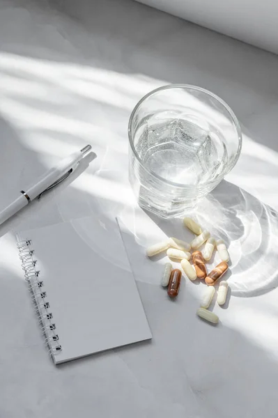 Healthy nutrition and diet planning concept, different supplement and vitamin pills and capsule, glass with pure water, pen and blank notebook mockup on white background, lifestyle sunlight shadow