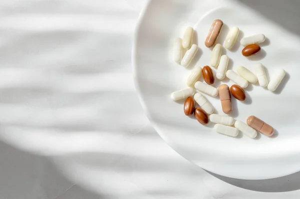 Minimal lifestyle aesthetic medicine concept. White and brown pills and capsules on white plate and marble table background with natural sun light shadows.