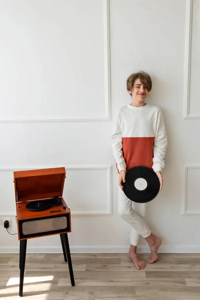 Retro vinyl record music trend aesthetic. Handsome teenager boy standing near wooden brown turntable, holding vinyl record in hands and smiling. Home interior, white wall background.