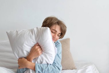 Günaydın, boş zaman eğlencesi, gençlik günlük yaşam tarzı konsepti. Genç çocuk yastığı kucaklıyor, yatakta oturuyor ve gülümsüyor. Boş beyaz duvar arkaplanı, minimalist kuzeyli yatak odası.