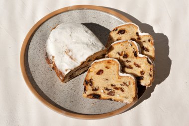 Christstollen, geleneksel Alman pastanesi. Meyveli ekmek ya da kek, kuru üzüm ve şeker tozu tabakta, bej bir masa üzerinde, güneş ışığında..