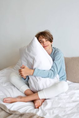 Yatakta oturup yastığı kucaklayan yakışıklı, samimi bir genç çocuk. Günaydın, sağlıklı günlük uyku konsepti, yaşam tarzı. Minimalist tarafsız yatak.