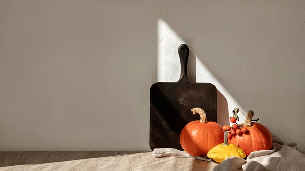 Orange pumpkins, wooden board, and linen tablecloth on beige table. Minimal kitchen autumn interior decor, empty wall background with natural sunlight shadow, fall vegetables harvest concept.