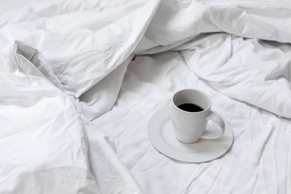 Cup with coffee drink standing on unmade bed with messy crumpled blanket, sheet and pillow. Lifestyle morning concept, coffee in bed.