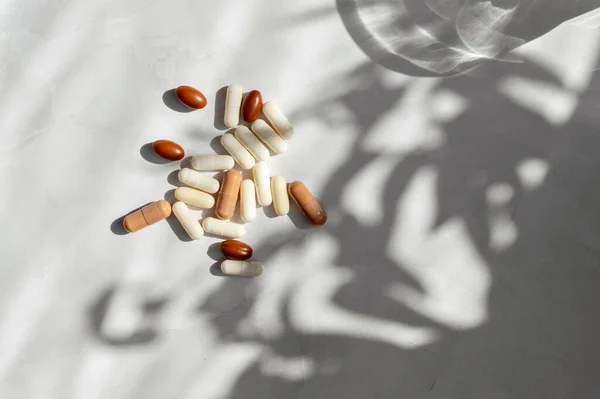 Brown and white supplement and vitamin capsules on white marble background with floral sunlight shadows
