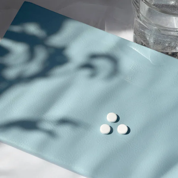 Healthcare minimal lifestyle concept. White pills on blue notebook with aesthetic sunlight shadows.