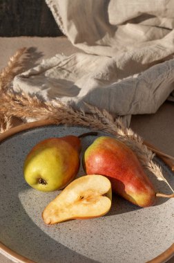 Olgun kırmızı ve sarı bütün ve yarım armut tabakta, keten masa örtüsü, bej masa arkasında kurutulmuş çayır çimeni güneş ışığında gölgelerle. Estetik yaşam tarzı meyvesini düşürür..