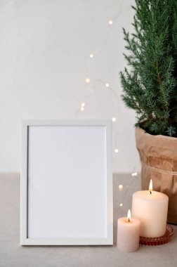 Boş resim çerçevesi maketi, mumlar, tencerede ardıç, tarafsız bej masa ve beyaz duvar arka planında çözünmüş çelenk ışıkları. Noel tatili İskandinav ev içi, ticari marka şablonu.
