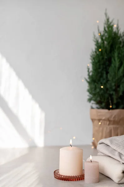 Scandinavian winter home interior decoration. Candles, juniper in pot, sweaters on beige table, empty white wall background with aesthetic sunlight shadows. Minimal white Christmas design template.