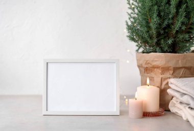 Masada boş beyaz çerçeve modeli, mumlar, tencerede ardıç, yün kazaklar, beyaz duvar arkası. Kışlık ev dekorasyonu, Noel ışıkları, estetik tatil şablonu..
