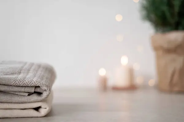 Wool sweaters stack on table on foreground. Defocused blurred candle lights and juniper in pot on background. Aesthetic minimal winter home interior backdrop.