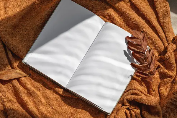 Blank open notebook mockup with empty sheets, brown fall leaves on terracotta knitted fabric background with natural sunlight shadows. Lifestyle aesthetic business brand or writer blog template.
