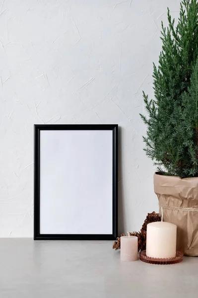 Blank picture frame mockup, candles, juniper in pot on neutral beige table, empty white wall background. Aesthetic minimalist winter home interior template, white Christmas decoration.