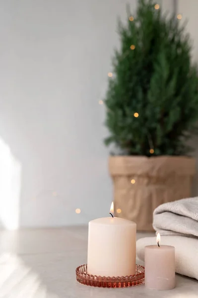 Minimalist aesthetic Christmas background with candle lights and blurred defocused juniper on table with garland lights. Empty neutral white wall background with sunlight shadows. Winter home decor.