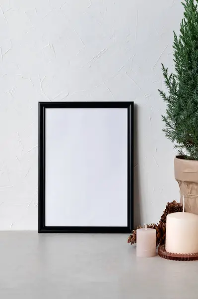 Minimalist aesthetic winter home interior design template. Black picture frame, poster mockup with white blank copy space, candles and pine tree in pot on beige table, neutral white wall background.